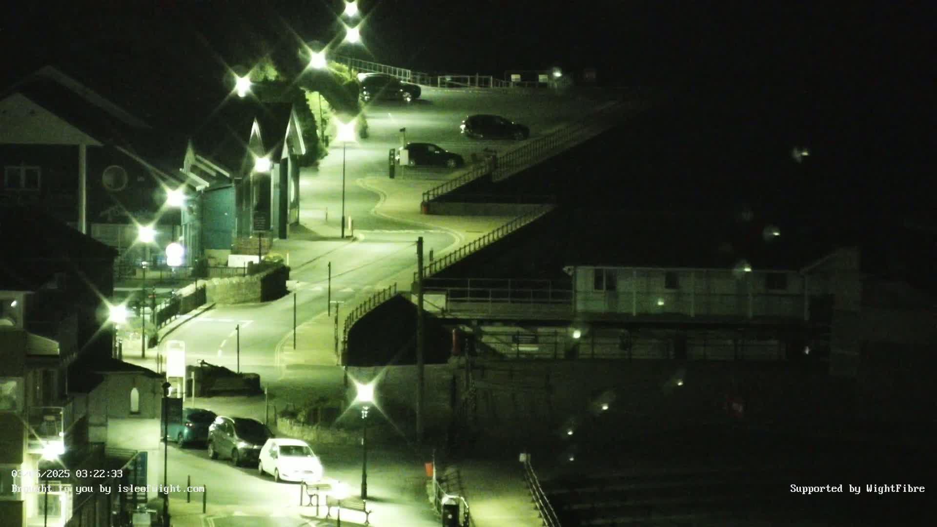 A nighttime aerial view shows a winding road illuminated by streetlights, with several cars parked in a lot and a few driving along the road, near some buildings.