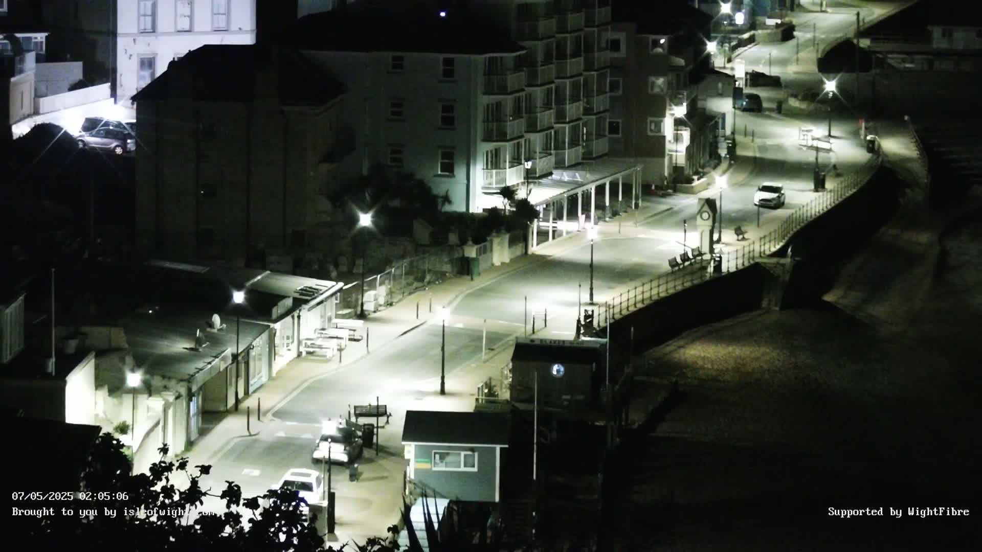 A nighttime, brightly lit street curves along a seaside town, lined with buildings and cars, next to a dark sandy beach.
