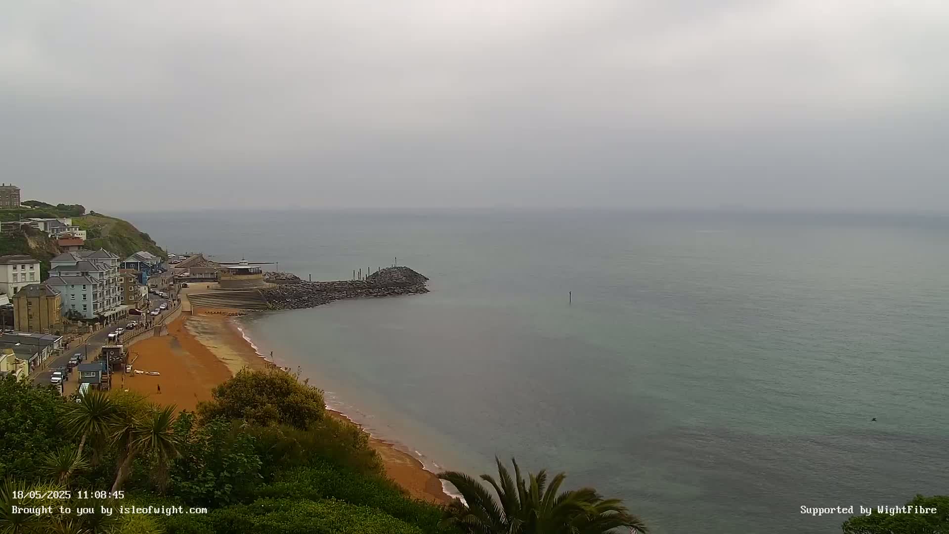 A coastal town overlooks a calm, grey sea and a sandy beach on an overcast day.