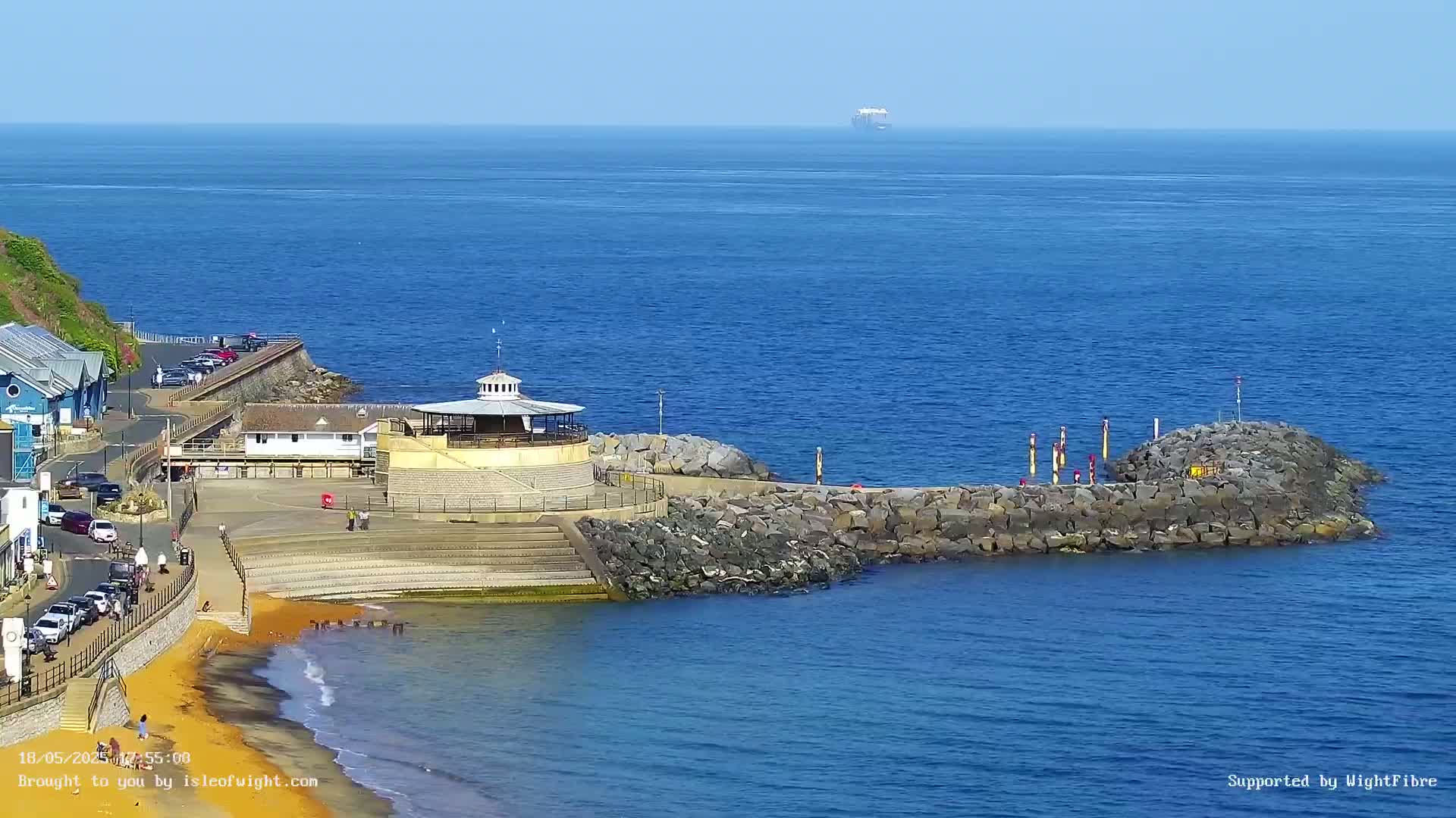 A seaside town scene on a sunny day shows a small beach, a stone breakwater, a gazebo-like structure, and a distant ship on a calm, blue ocean.