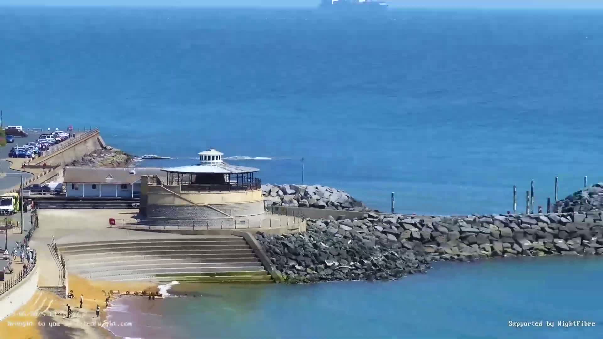 A seaside scene on a sunny day shows a round gazebo on a rocky breakwater, steps leading down to a small sandy beach with a few people, and a parking lot and buildings behind it.