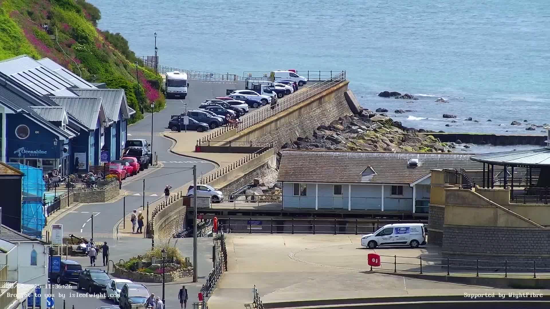A coastal town scene on a sunny day shows a waterfront with parked cars, buildings, and pedestrians, along with a rocky shoreline and calm ocean water.
