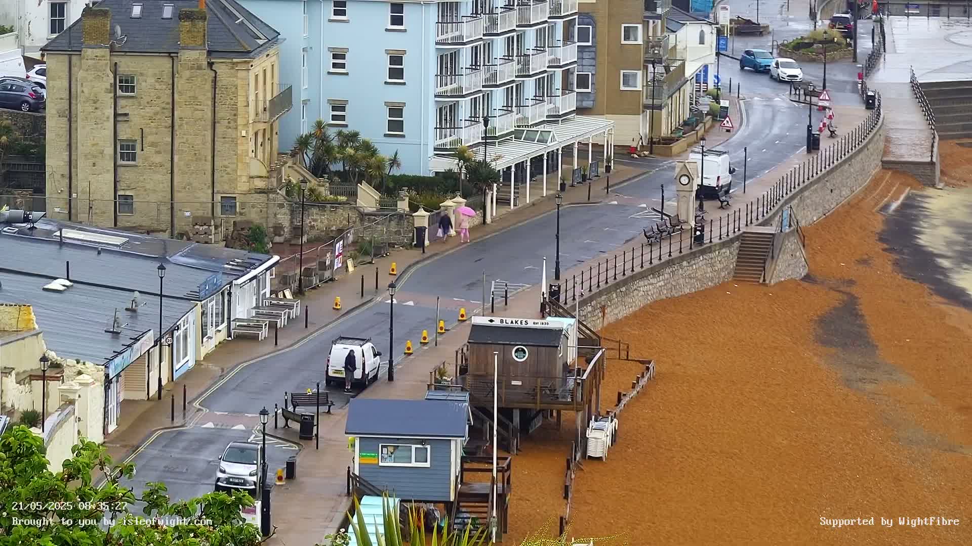 A rain-slicked seaside street scene shows buildings, parked cars, pedestrians, and a beach with reddish-brown sand.