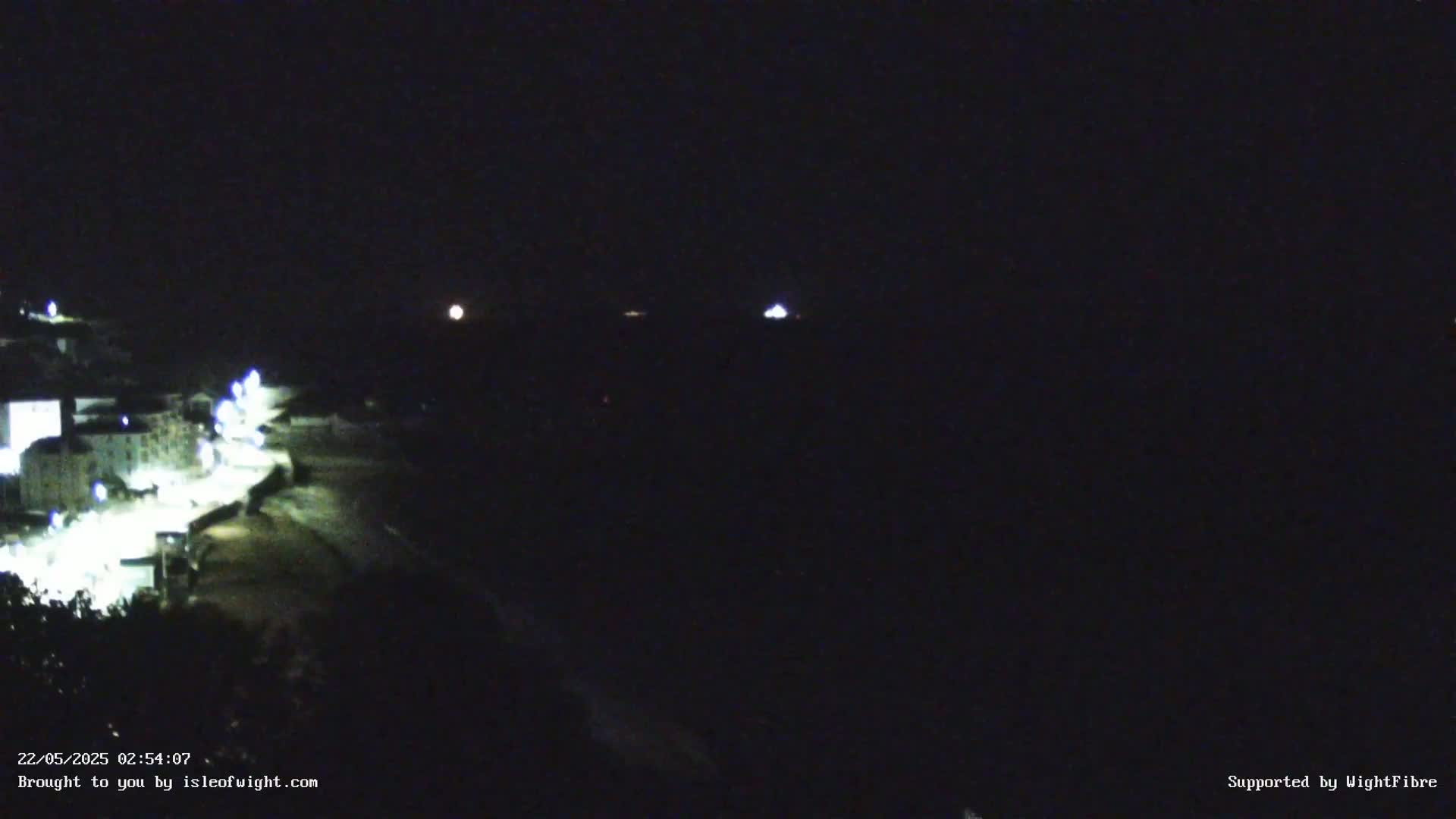 A nighttime, long-range view shows a moonlit coastline with a town's lights visible on a dark, seemingly calm body of water.