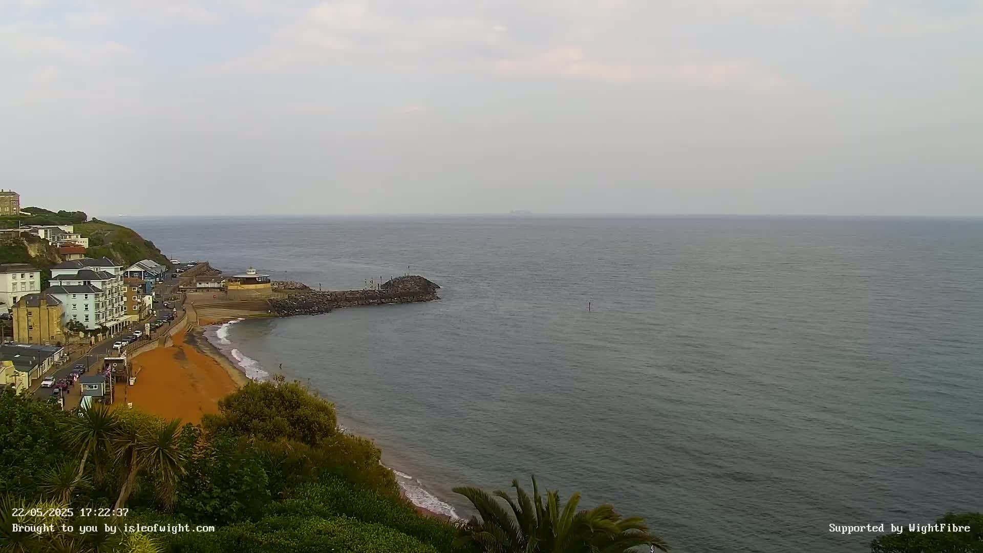 A coastal town with buildings and a sandy beach is seen overlooking a calm, gray sea under an overcast sky.