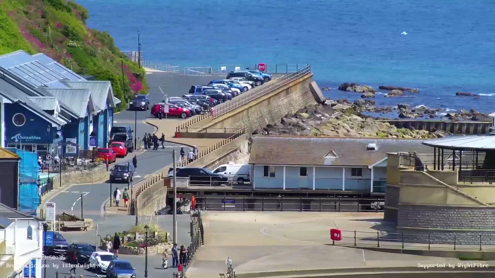 A seaside town scene on a sunny day shows a parking lot next to a seawall with buildings and people walking along a road.