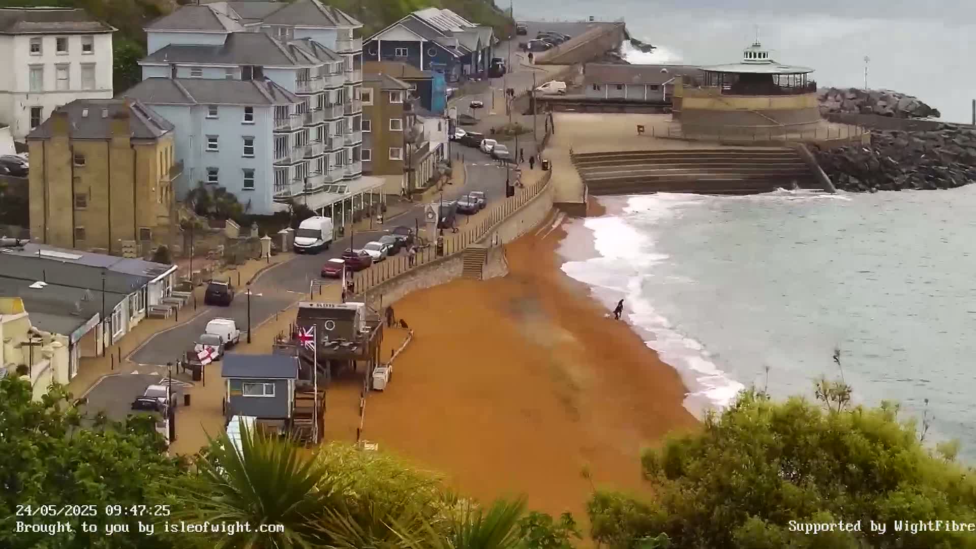 A red sand beach with small waves laps against a sea wall next to buildings and a roadway.