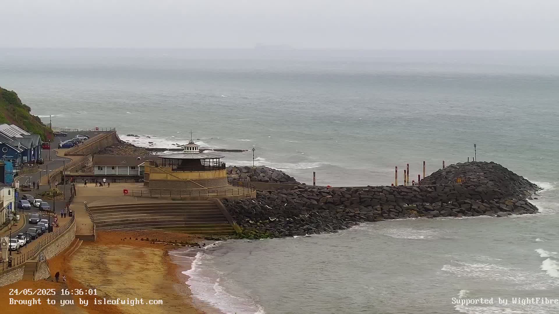 A rocky breakwater protects a sandy beach and coastal buildings from a gray, overcast sea with small waves.