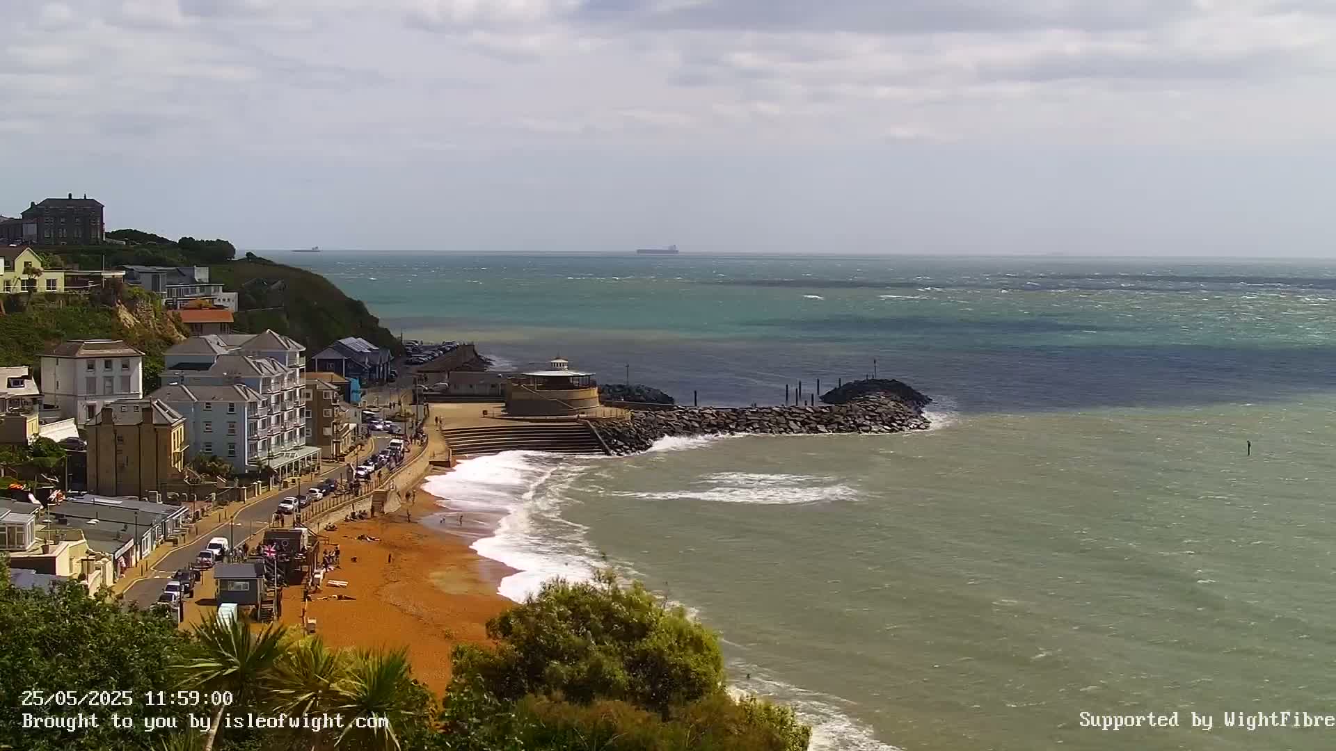 A seaside town with a sandy beach and buildings on a cliff overlooking a choppy sea under a partly cloudy sky.