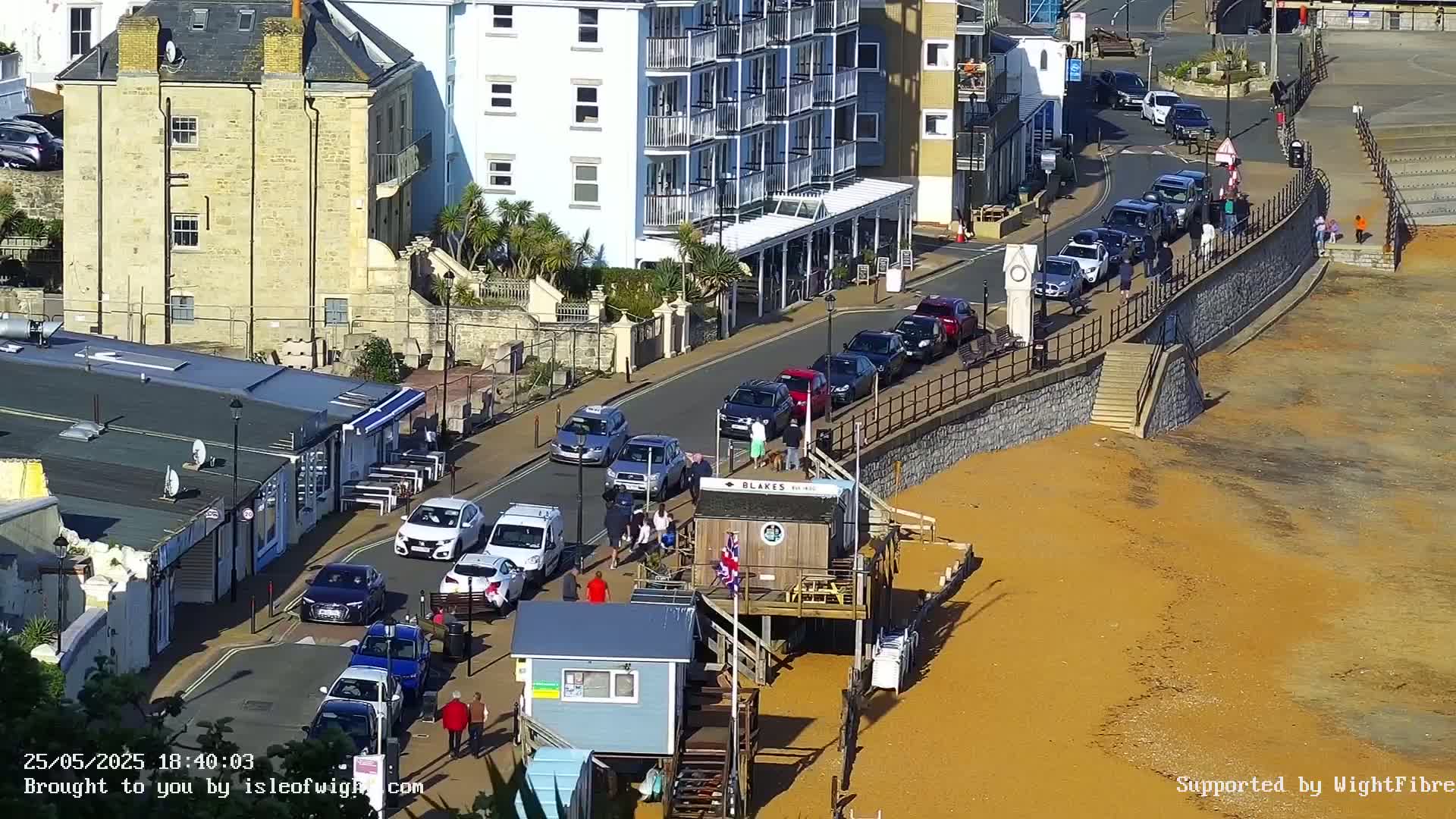 A sunny seaside town scene shows parked cars lining a street next to a sandy beach with a small beach hut and several buildings visible.