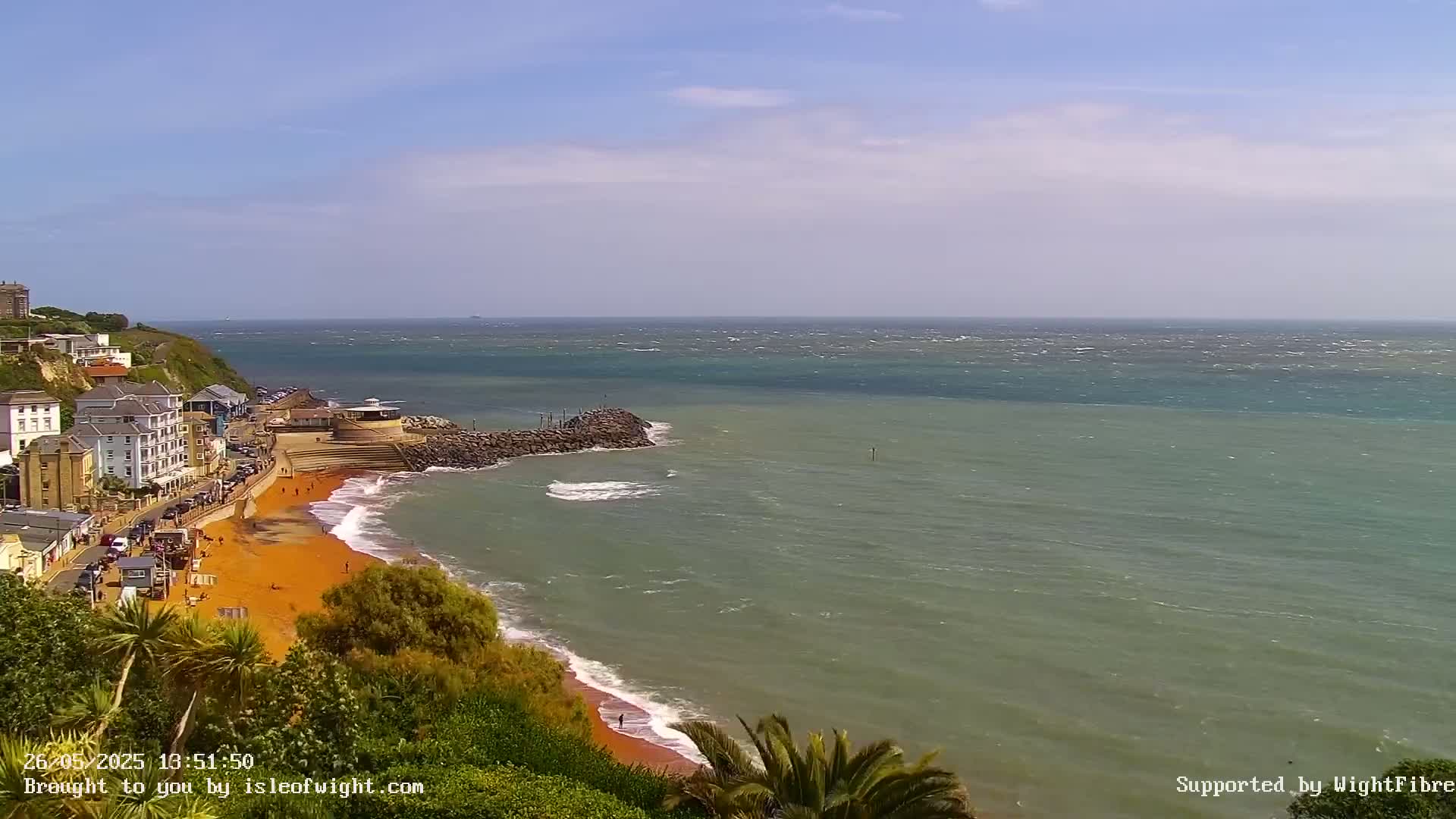 A seaside town with buildings on a cliff overlooking a windy ocean with a red-sand beach.