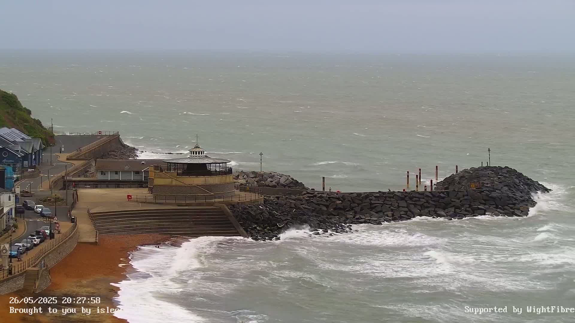 A rocky breakwater protects a red sand beach and seaside buildings from a choppy, gray sea under an overcast sky.