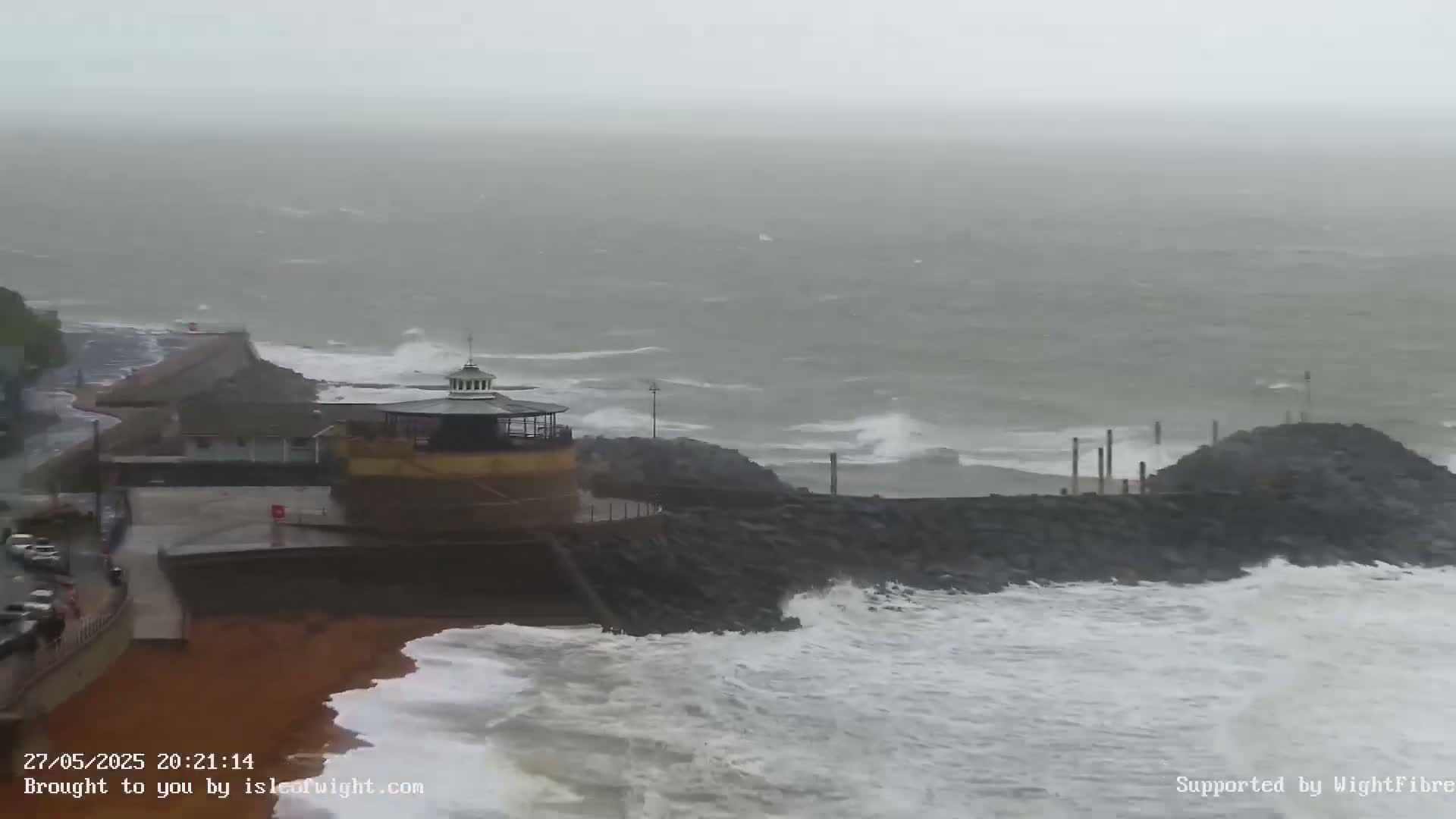 Large waves crash against a rocky shoreline near a small building with a dome-shaped roof during stormy weather.