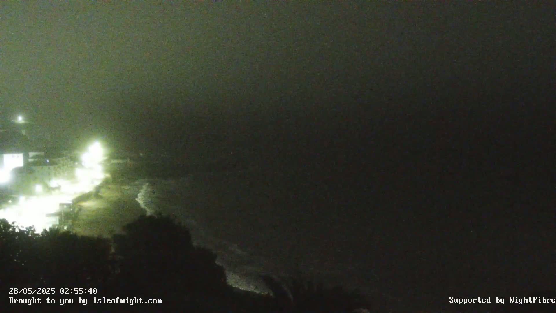 A nighttime, hazy view of a coastline shows dimly lit buildings and a dark beach with gentle waves.