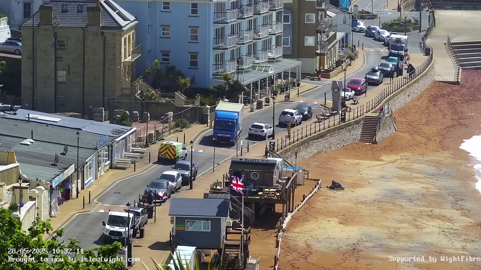A seaside street scene on a sunny day shows a line of parked cars, several service vehicles, and a beach with a reddish-brown sand.