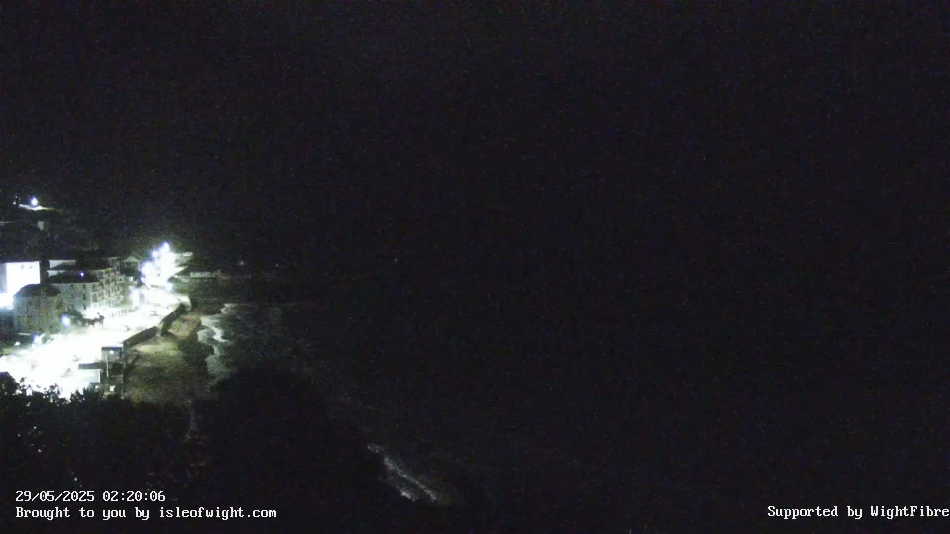 A nighttime coastal scene shows a dimly lit town on a hillside next to a dark ocean with waves.