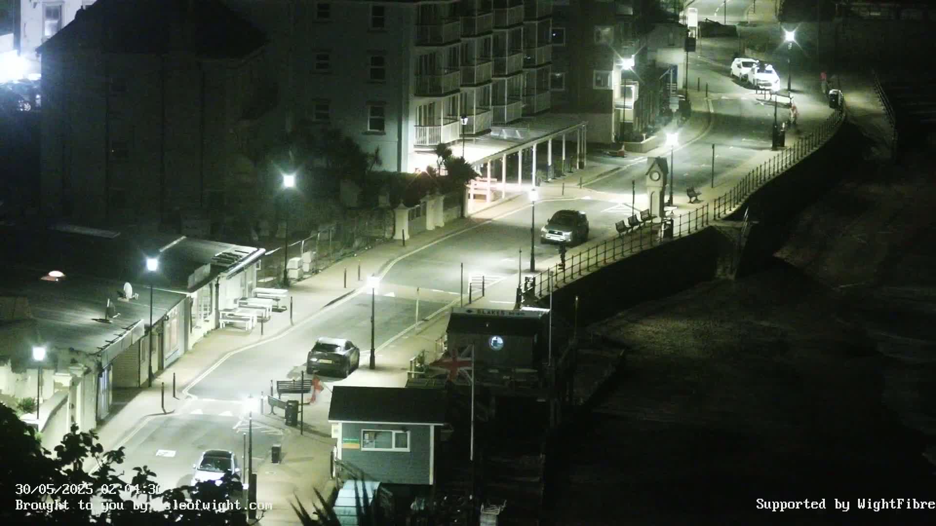 A nighttime, high-angle view of a seaside street with cars parked along the road, buildings lit by streetlights, and a dark beach below.