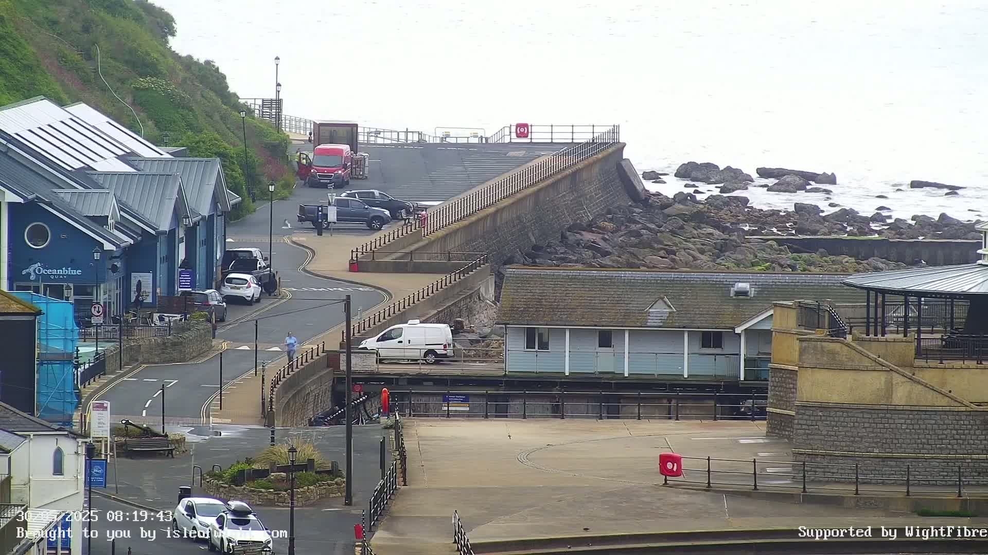 A coastal town scene on an overcast day shows a road curving past buildings and a seawall overlooking rocky shoreline and the ocean.