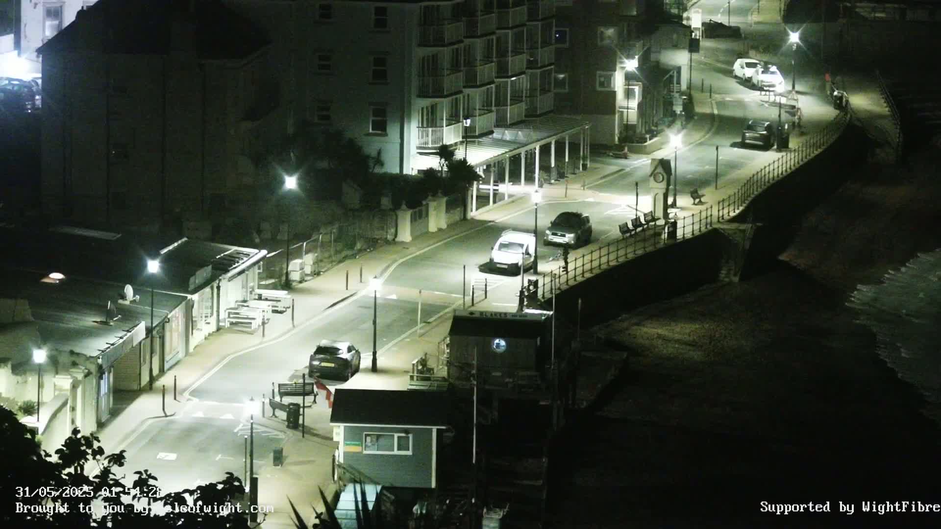 A nighttime, high-angle view of a street with several cars parked along a seawall, and buildings illuminated by streetlights.