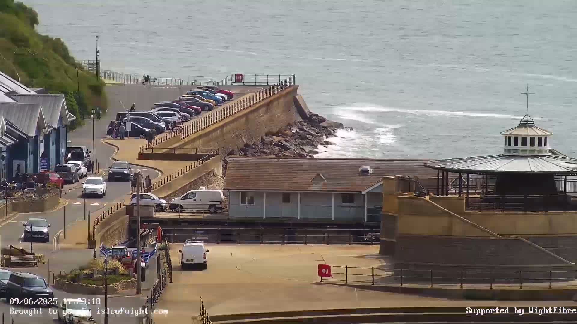 A seaside scene with a parking lot, buildings, and a gazebo overlooking a rocky shore with gentle waves on a sunny day.
