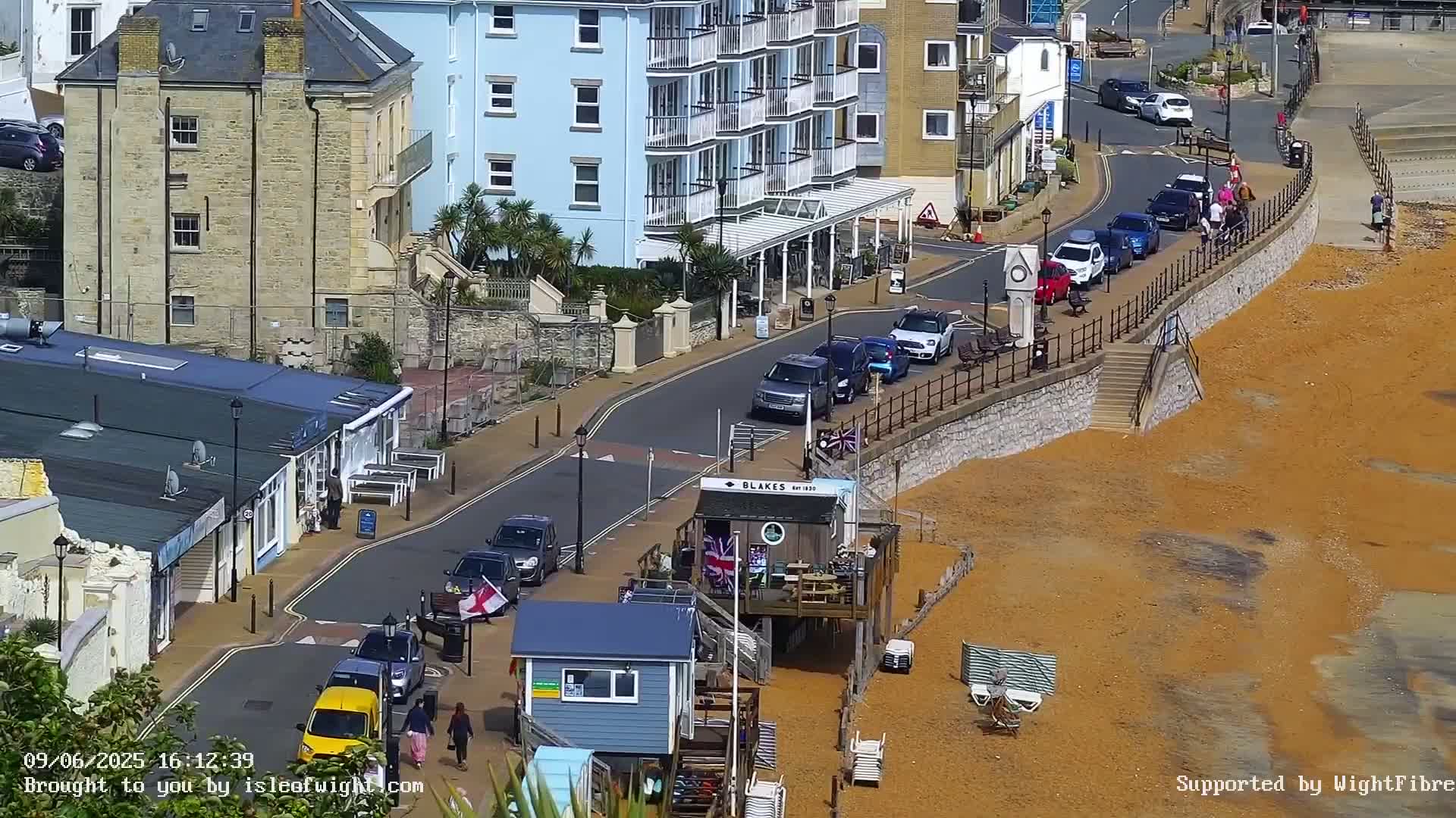 A sunny seaside town scene shows a street with parked cars, pedestrians, and buildings, leading down to a sandy beach with beach chairs and small structures.