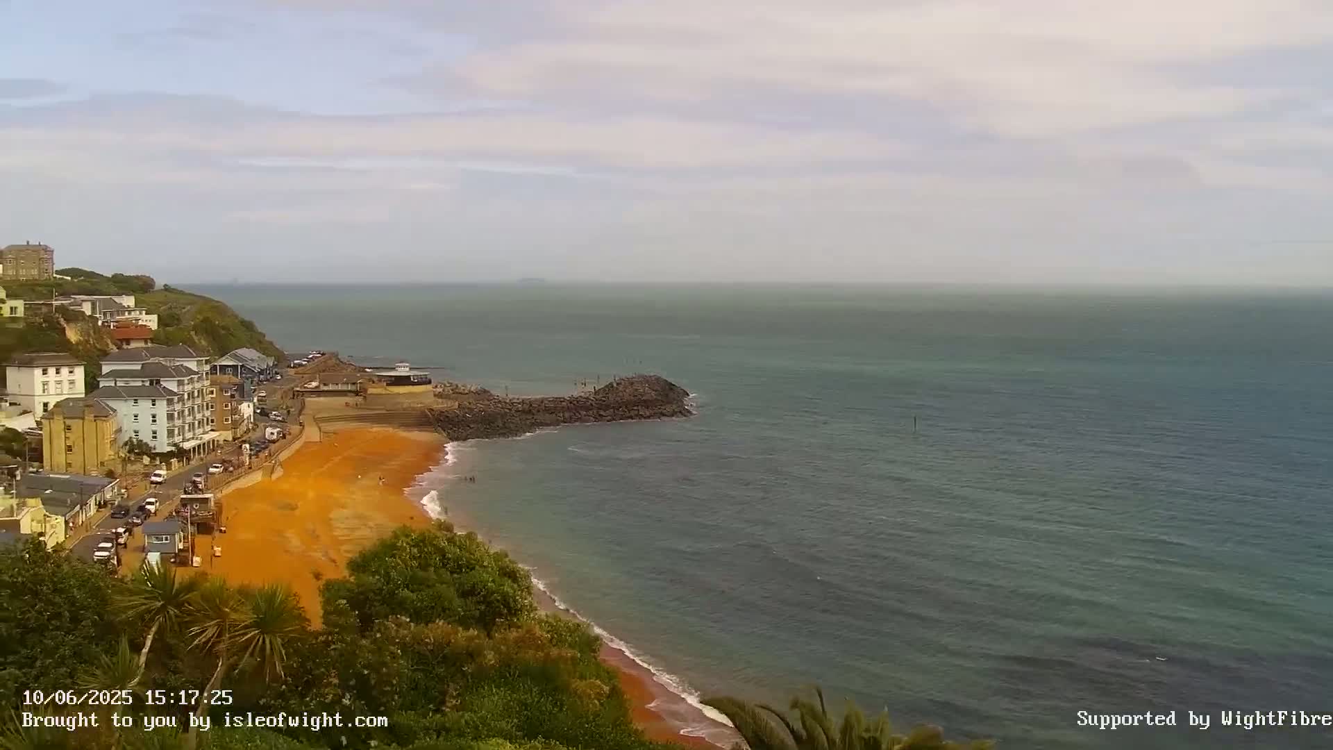 A red-sand beach curves along a coastline, bordered by buildings and green vegetation, under an overcast sky with calm, dark-blue water extending to the horizon.