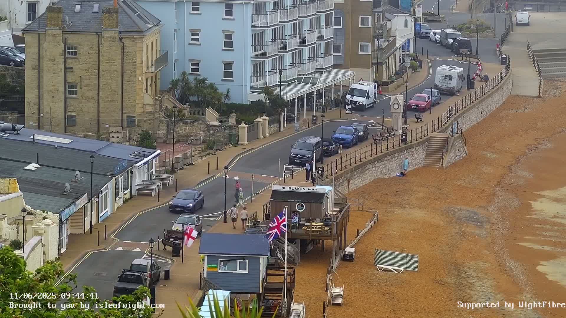 A street scene shows several cars parked along a road next to a beach with reddish-brown sand under an overcast sky.