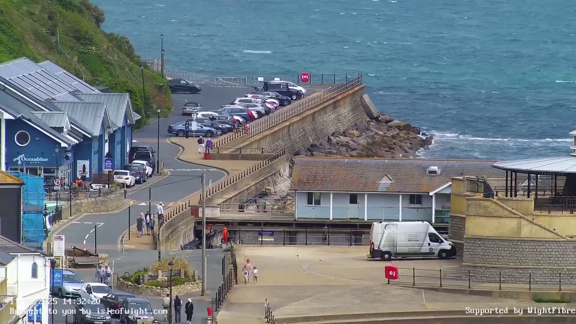 A seaside town scene on a partly cloudy day shows a parking lot, buildings, people walking, and a rocky coast with choppy water.