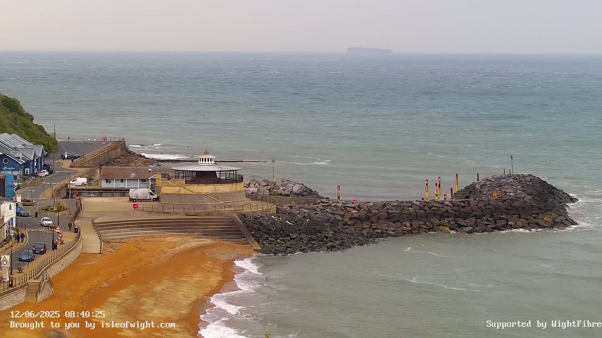 A rocky breakwater protects a sandy beach and a small, round bandstand overlooking a choppy sea under an overcast sky.