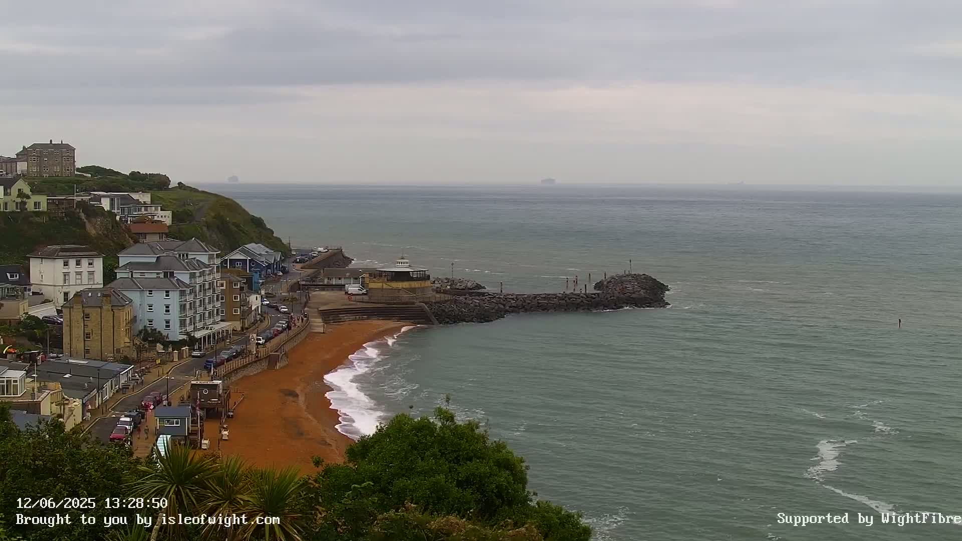 A coastal town with red sand beach and buildings on a cliff overlooks a calm, gray sea under an overcast sky.