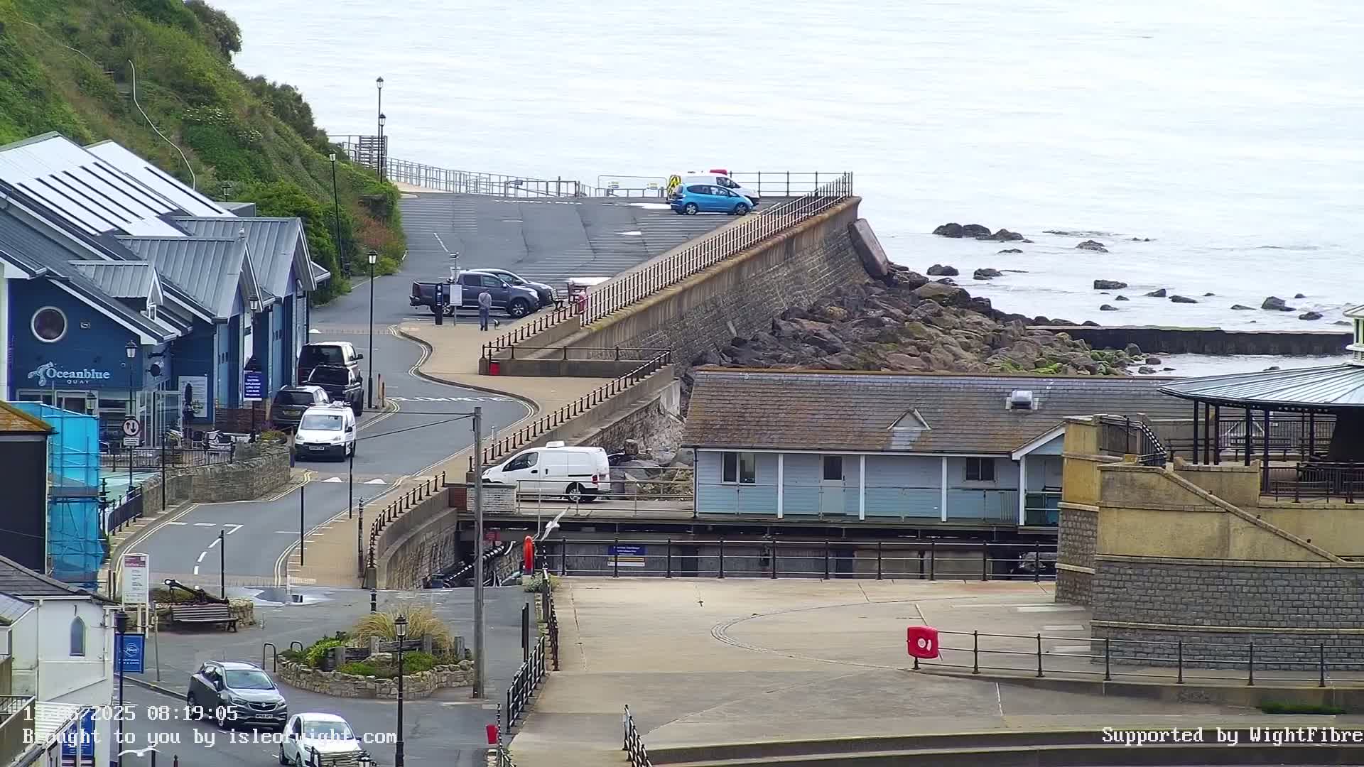 A coastal scene with overcast skies shows a parking area, buildings, and a roadway leading down to the rocky ocean shore.