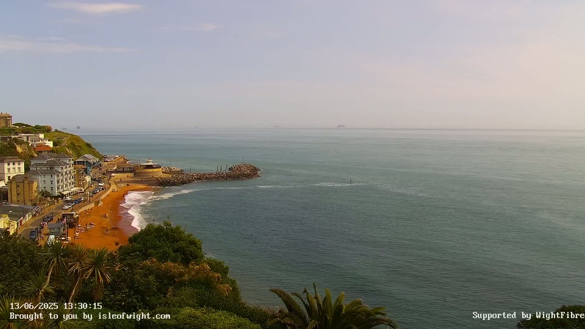 A hazy, sunny day overlooks a coastal town with a red sand beach and calm ocean stretching to the horizon.