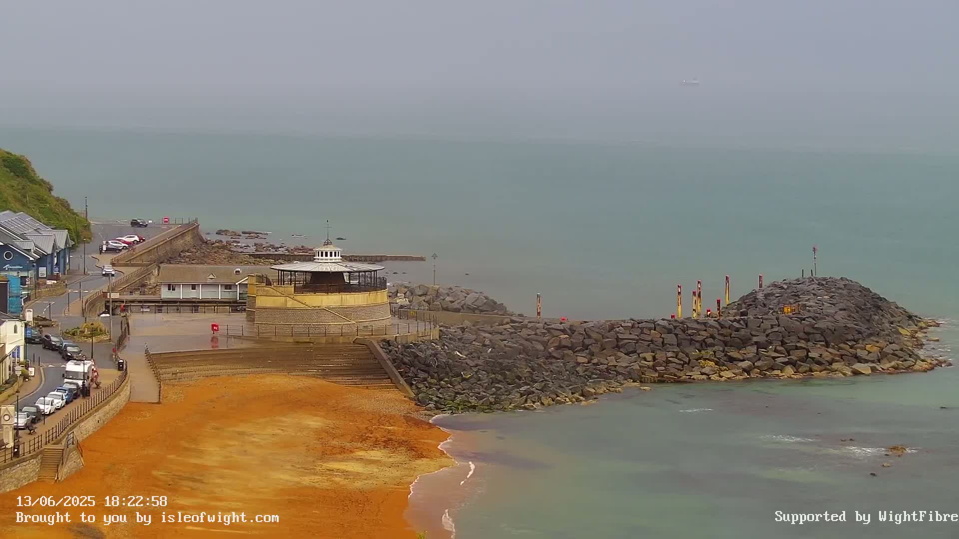 A coastal scene on an overcast day shows a rocky breakwater jutting into a calm sea, next to a sandy beach with a small building and a parking area.