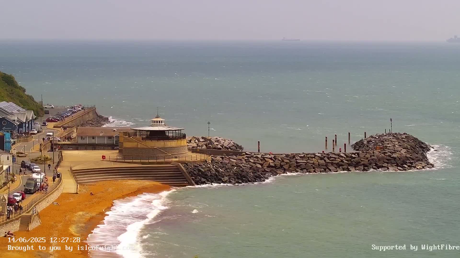 A seaside town with a small, round gazebo on a rocky jetty, a sandy beach, and a calm sea with a slight wind.