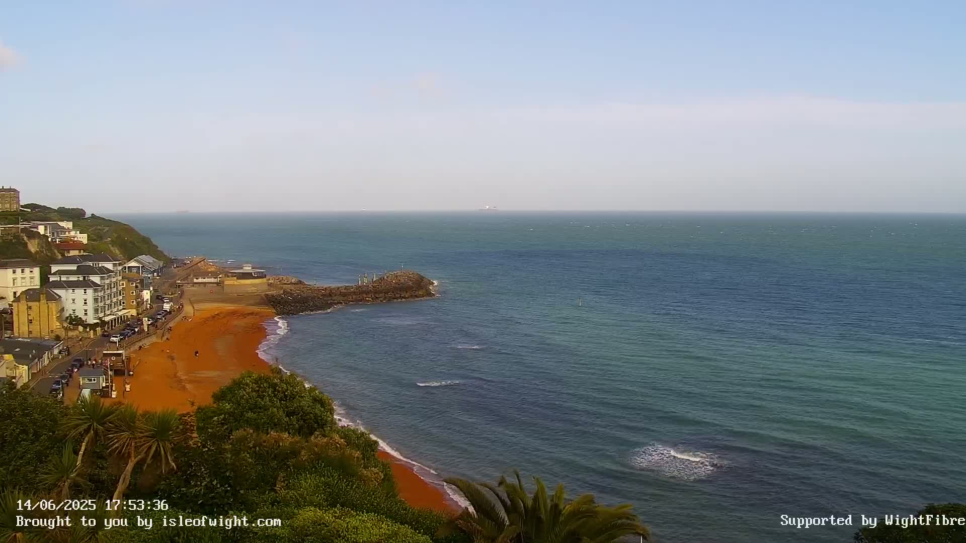 A coastal town overlooks a calm, teal ocean with a reddish-orange sand beach.