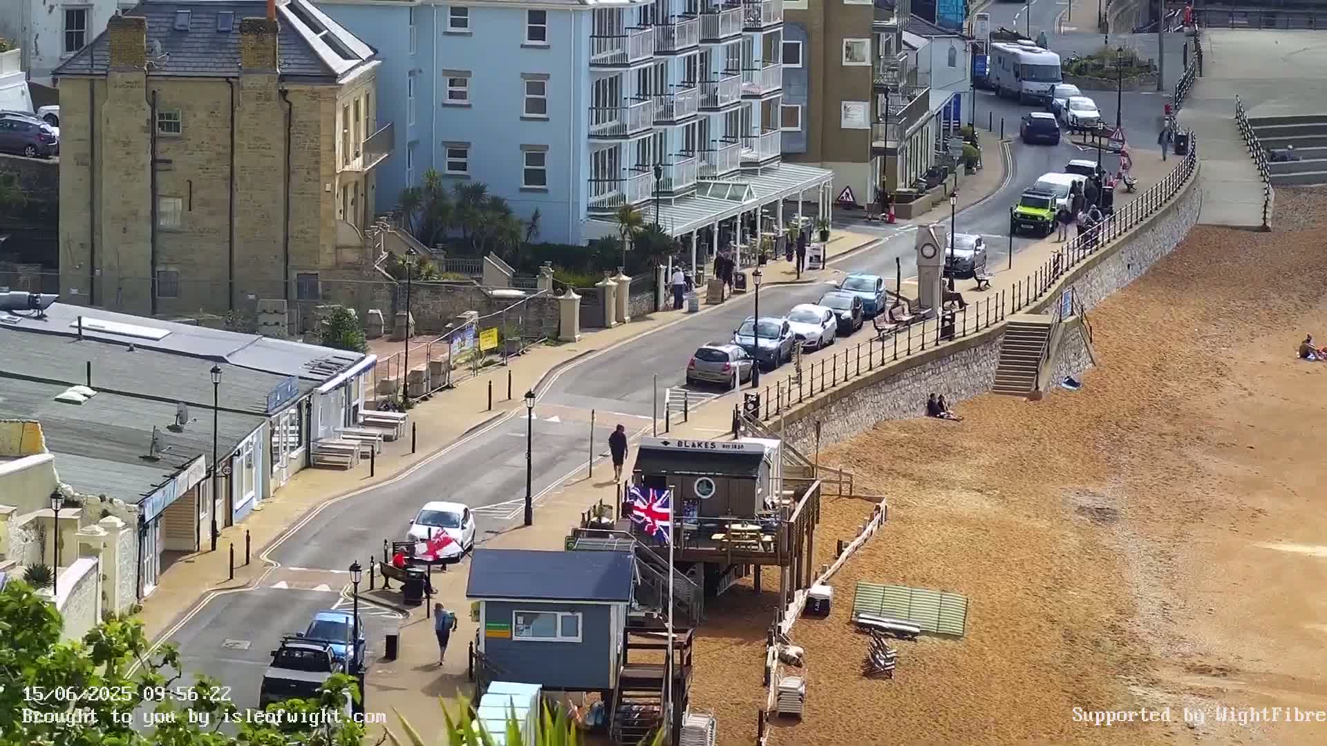 A sunny seaside town scene shows a street with parked cars, pedestrians, and buildings, leading down to a sandy beach with a few people relaxing.