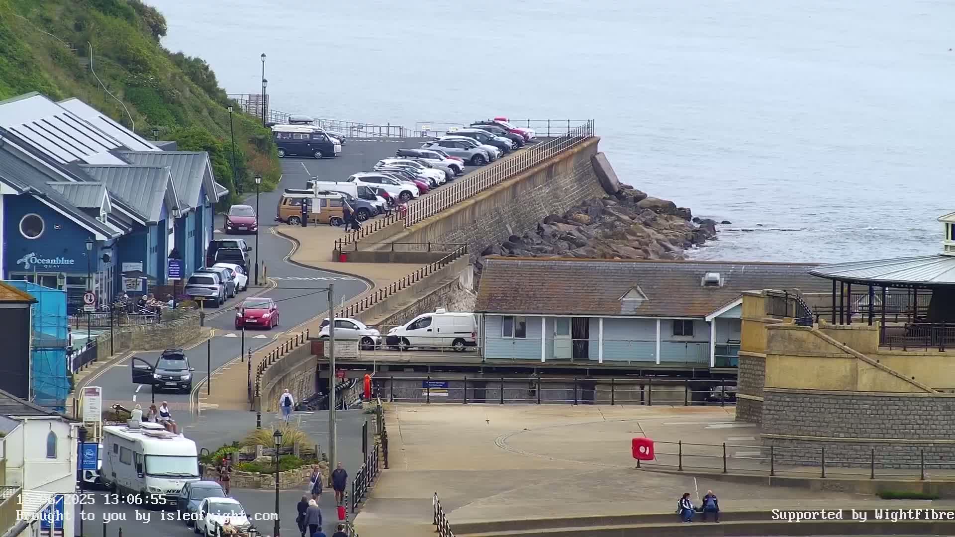 A seaside town scene shows a parking lot and buildings along a rocky coast under an overcast sky.