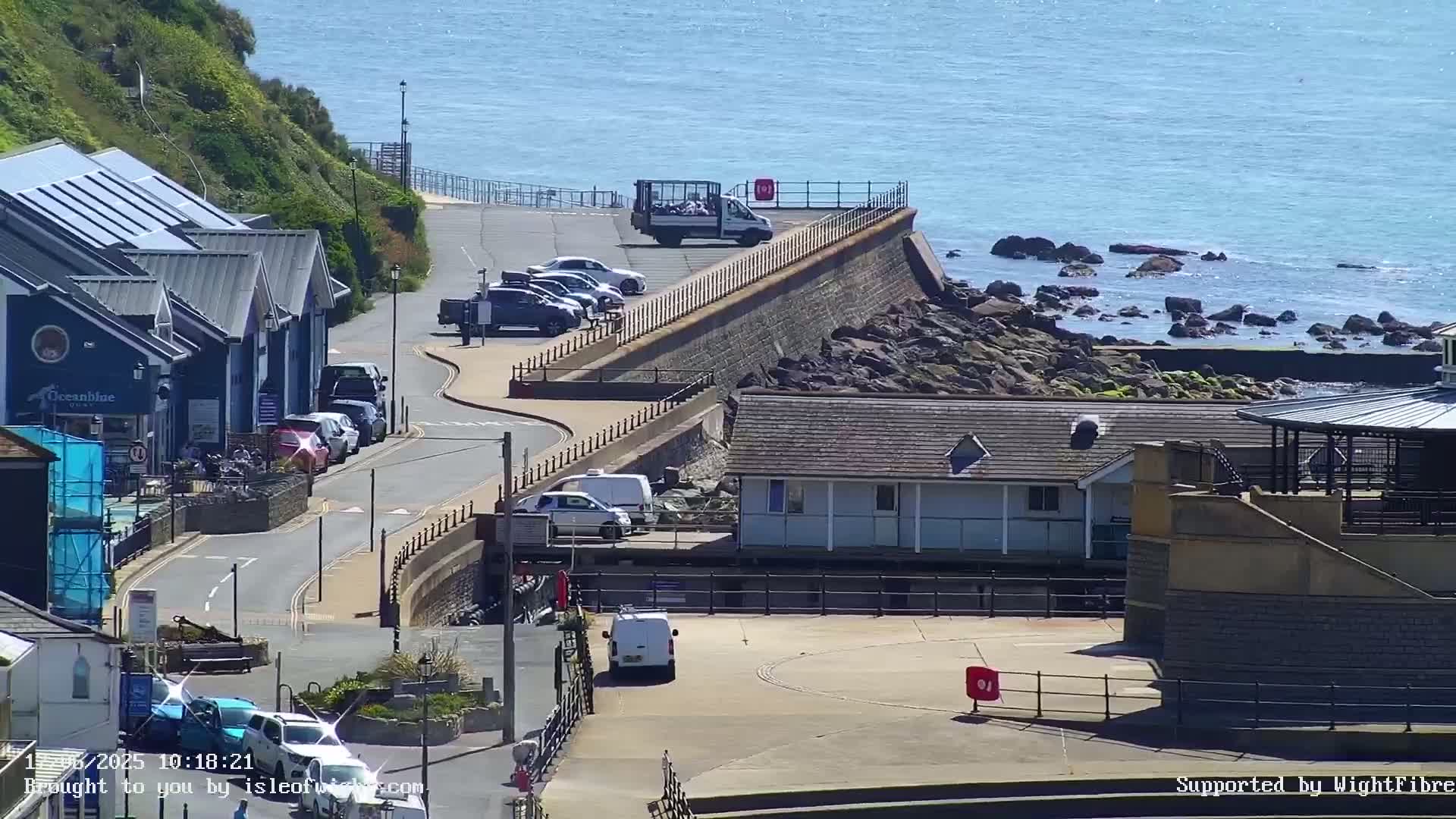 A coastal scene on a sunny day shows a parking area next to a sea wall with several cars, vans, and a building along a curving road.