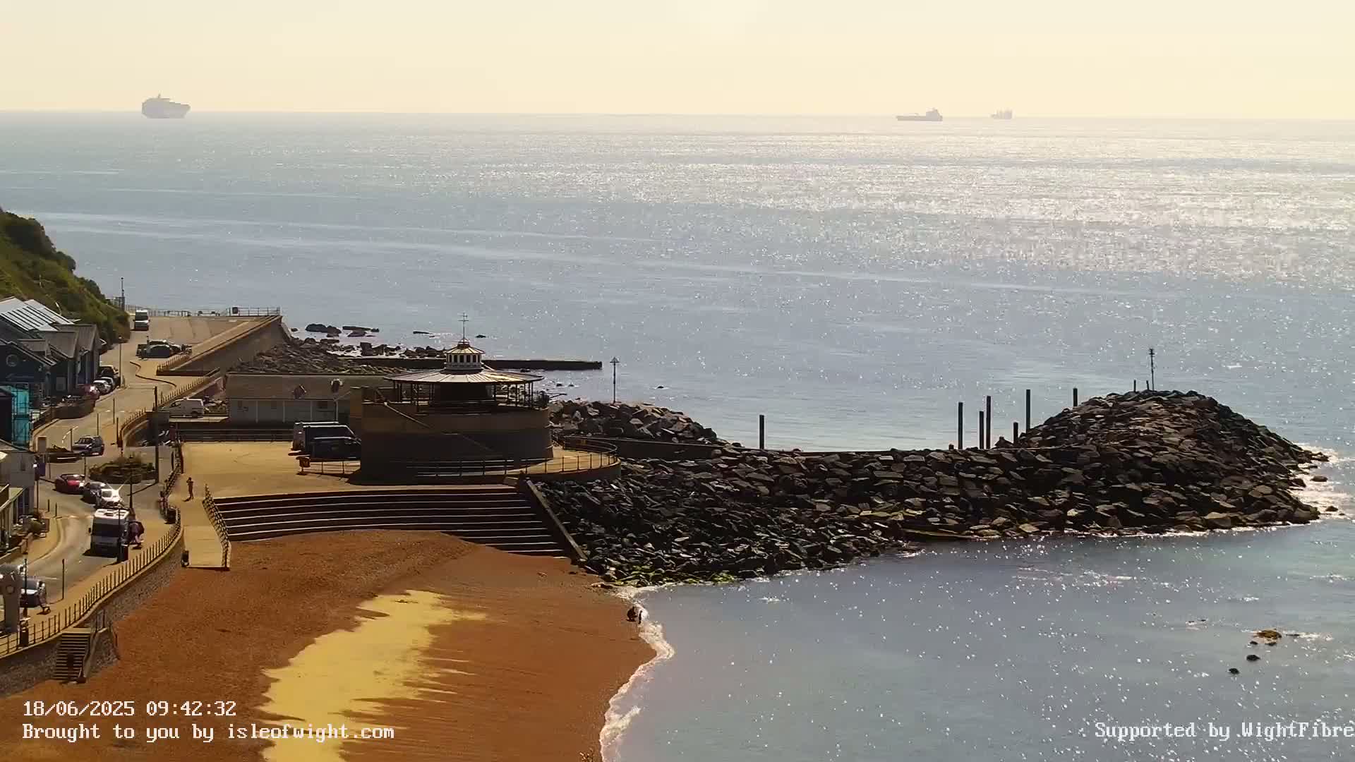 A coastal scene on a sunny day shows a sandy beach, a rocky breakwater, a small gazebo, and distant ships on a sparkling sea.
