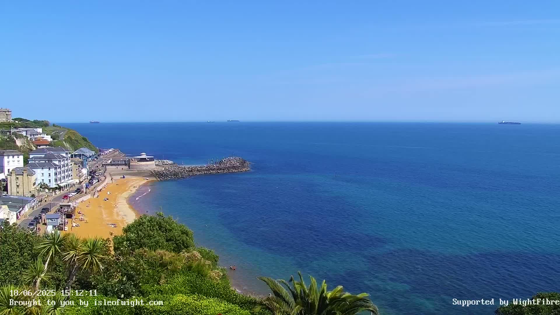 A sandy beach with buildings and vegetation overlooking a calm, blue ocean on a sunny day.