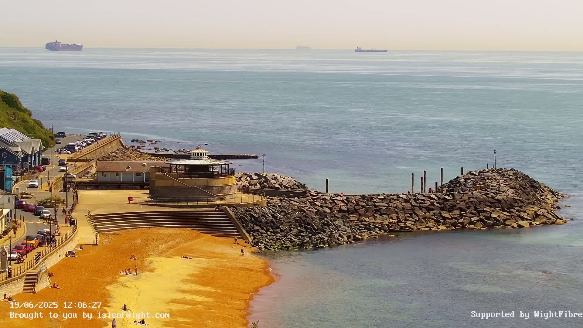 A sandy beach with a few people on it, a stone breakwater, a small building with a cupola, and several parked cars, all overlooking a calm sea with distant ships on a sunny day.