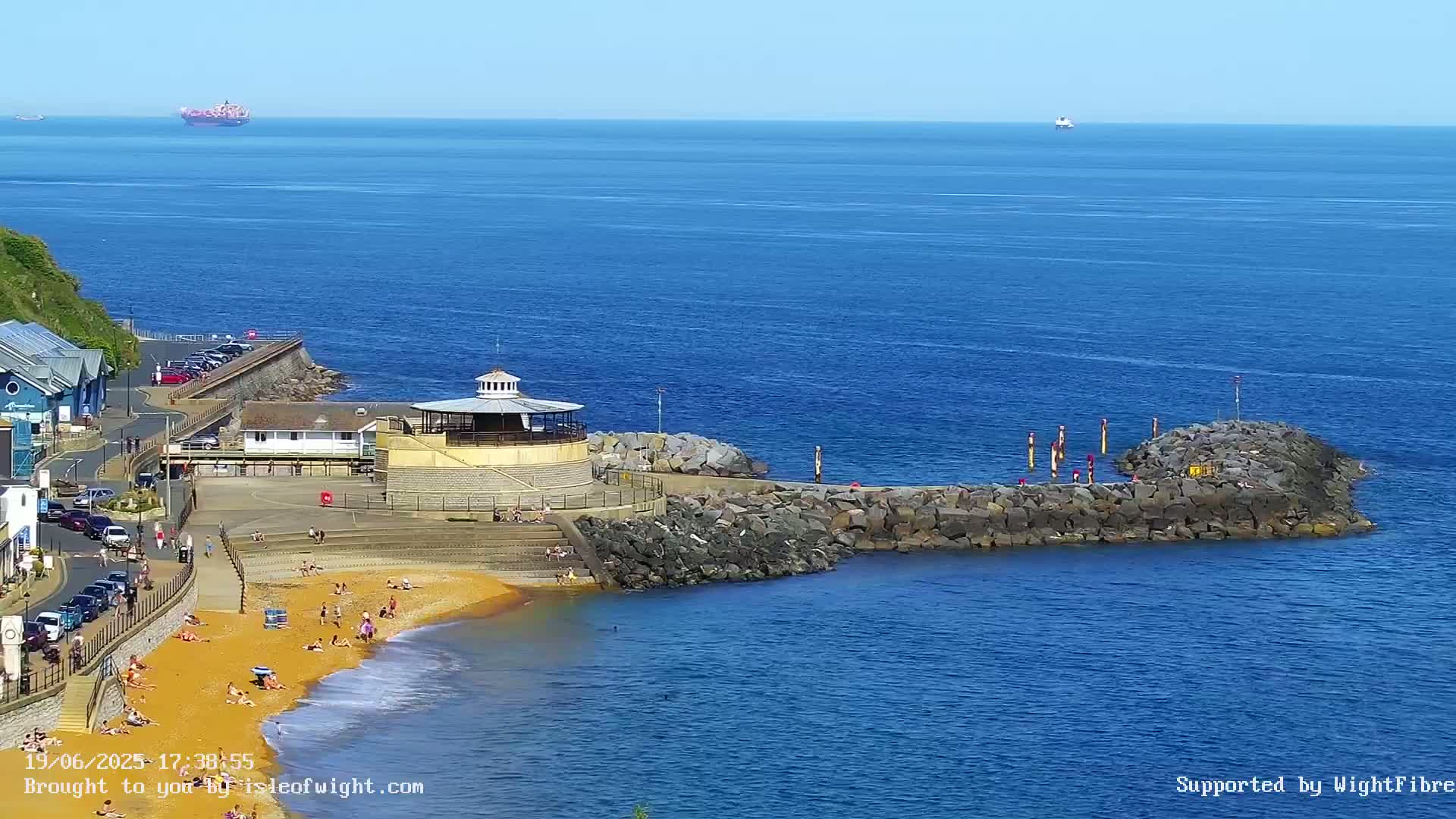 A sandy beach with people sunbathing and swimming is nestled beside a rocky breakwater and a small, round building, all under a clear, sunny sky, with a calm ocean extending to the horizon.
