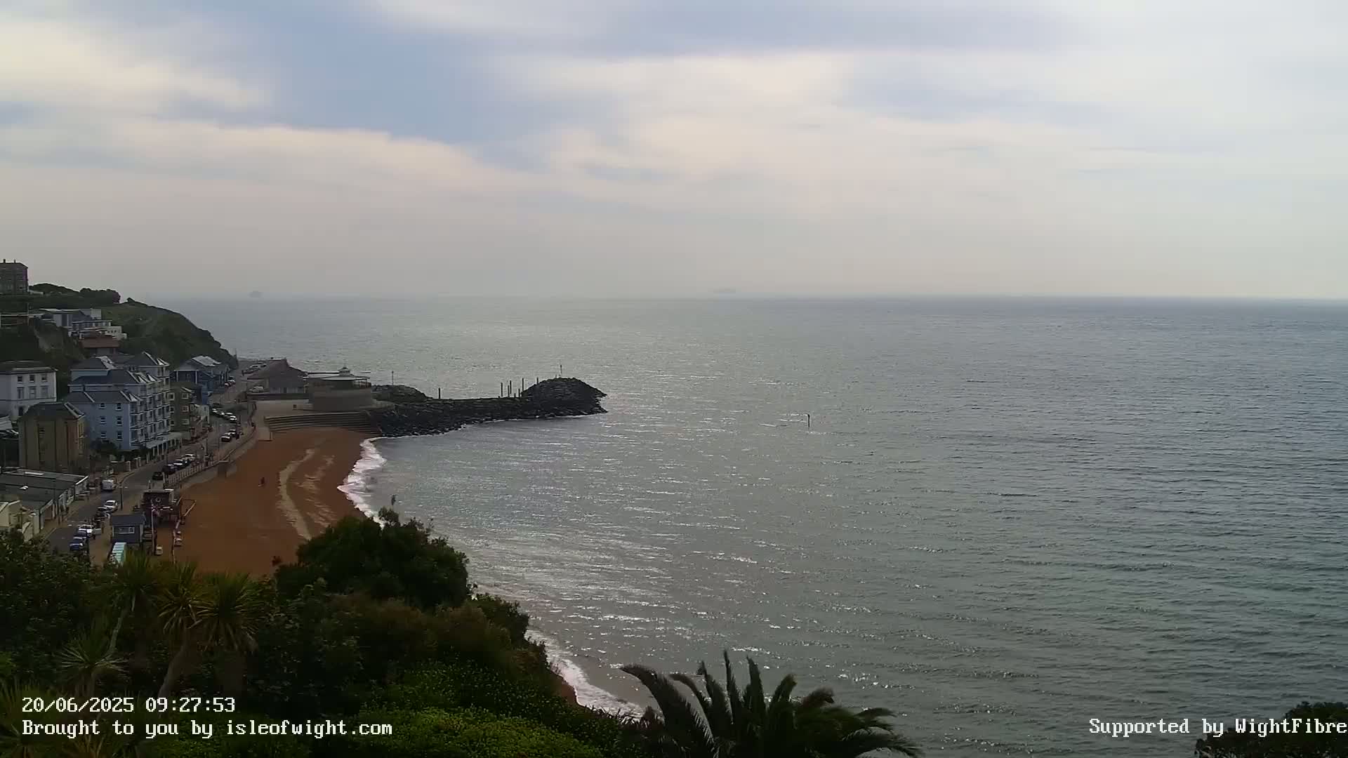 A sandy beach with buildings on a cliff overlooking a calm, gray sea under a mostly cloudy sky.