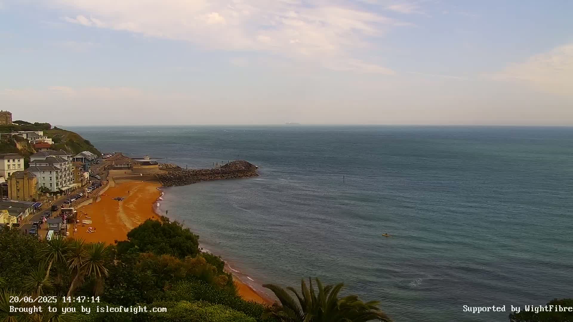 A partly cloudy day shows a coastal town overlooking an orange sand beach and calm blue ocean with a single person in a kayak.