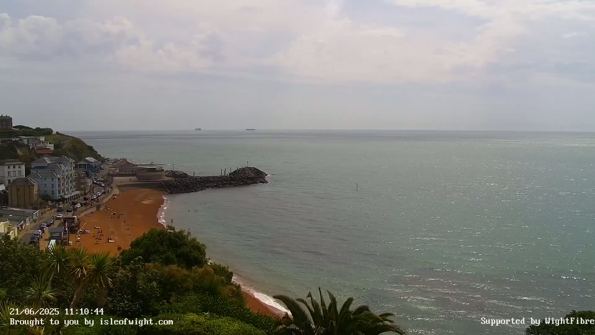 A mostly sunny day overlooking a red sand beach with buildings and a calm sea.