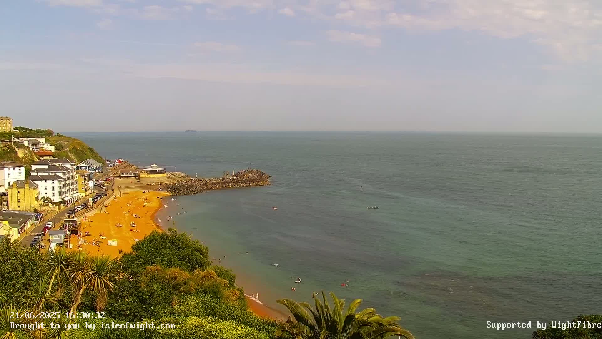 A sunny day shows a crowded sandy beach curving into a calm sea, next to buildings and green vegetation on a cliffside.