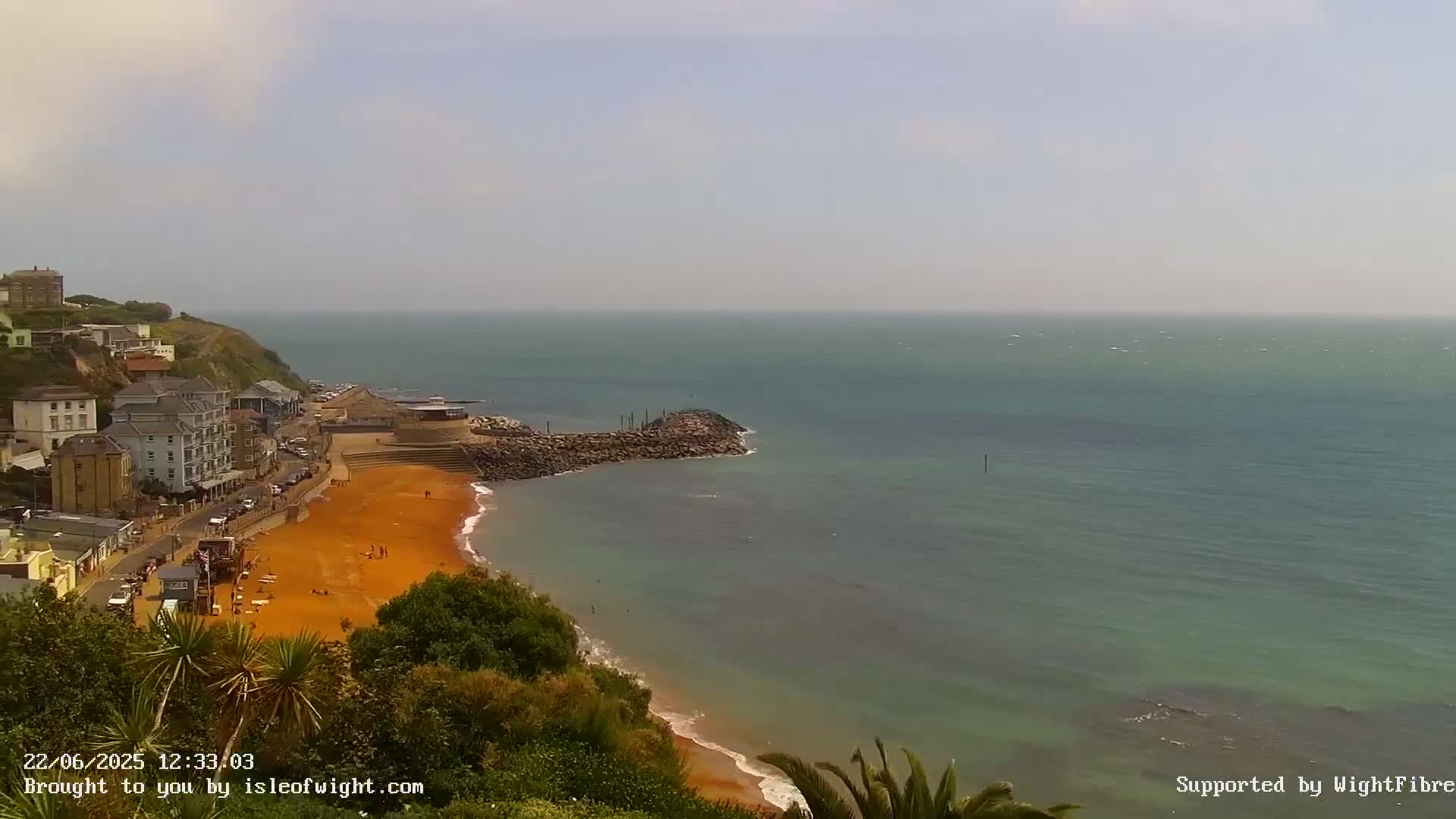 A hazy day reveals a coastal town nestled on a hillside overlooking an orange-sand beach and calm, teal-colored ocean.