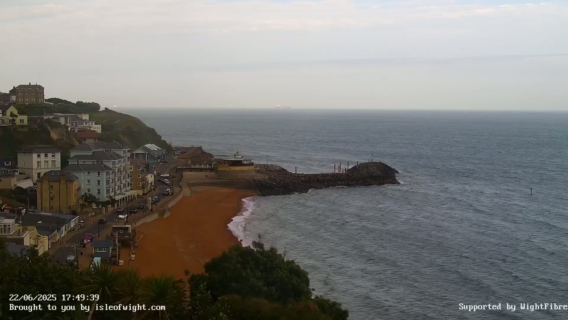 A coastal town overlooks a red sand beach and a calm, gray sea on an overcast day.