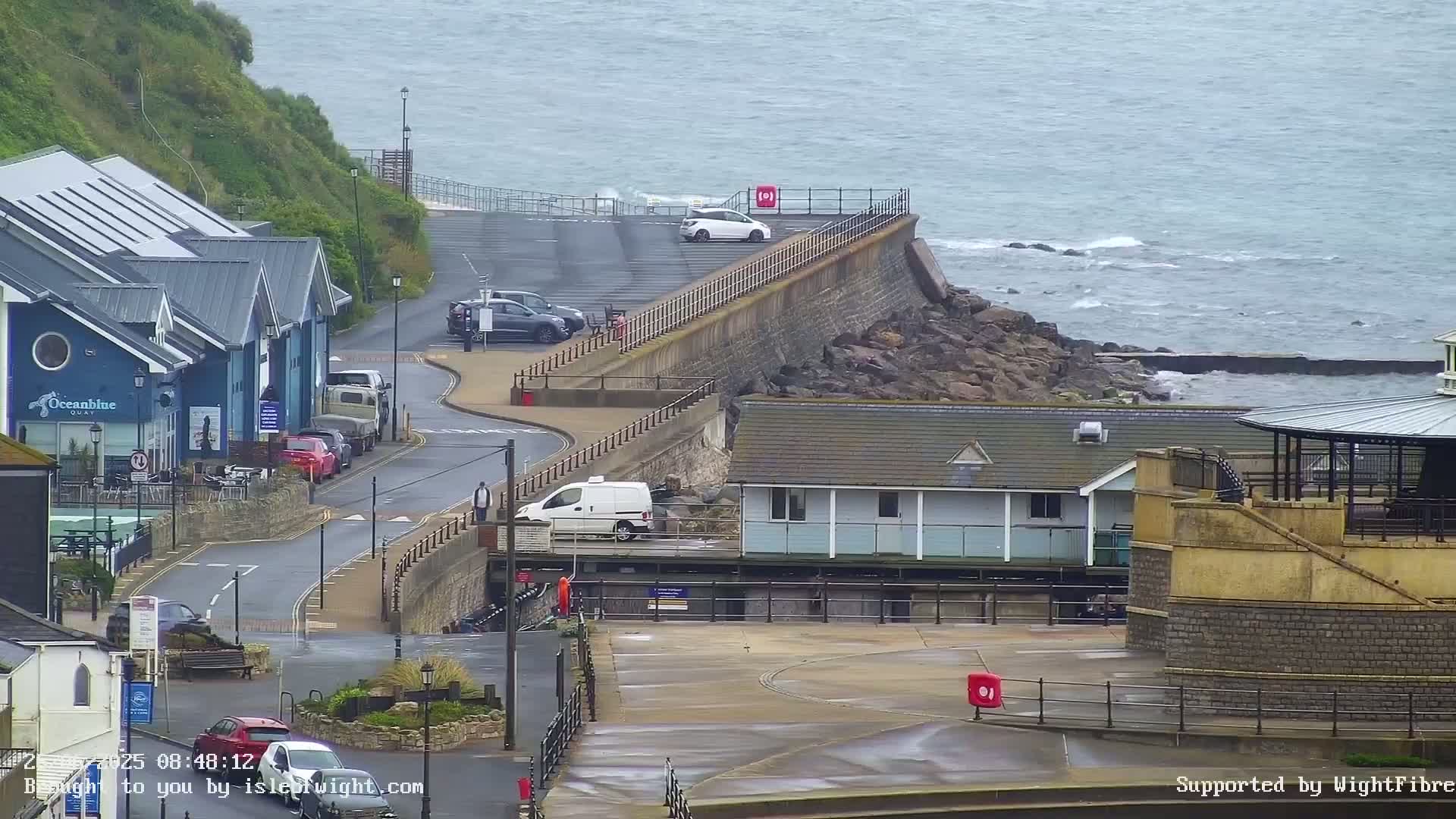 A coastal scene on an overcast day shows a small parking area next to a sea wall, several buildings, and a few cars parked along a road.
