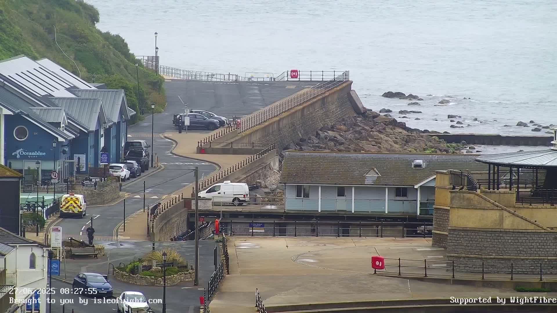 A coastal scene on an overcast day shows a road curving past buildings and a seawall that protects a rocky shoreline and calm ocean.